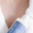Mão segurando o sabonete facial Soft Clean Sousènsi, frasco com válvula pump, sobre roupão branco
